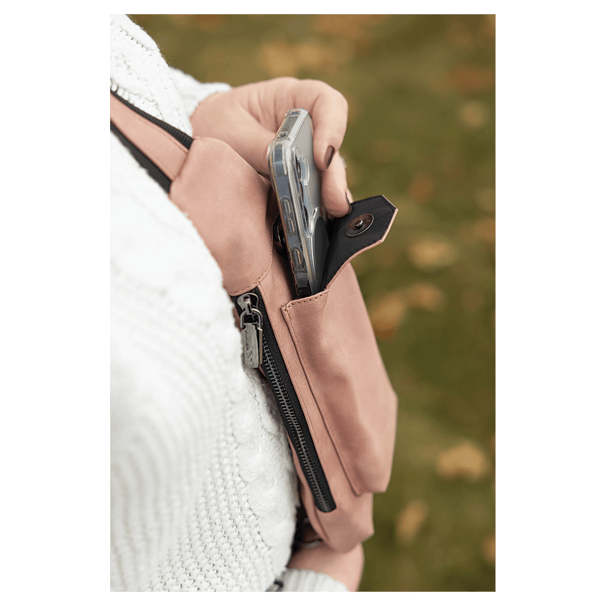A person in a white sweater removes a phone from a pink crossbody bag front snap pocket outdoors against autumn leaves.