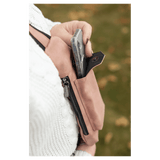 A person in a white sweater removes a phone from a pink crossbody bag front snap pocket outdoors against autumn leaves.