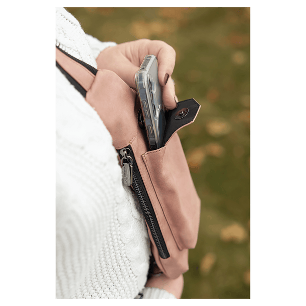 A person in a white sweater removes a phone from a pink crossbody bag front snap pocket outdoors against autumn leaves.