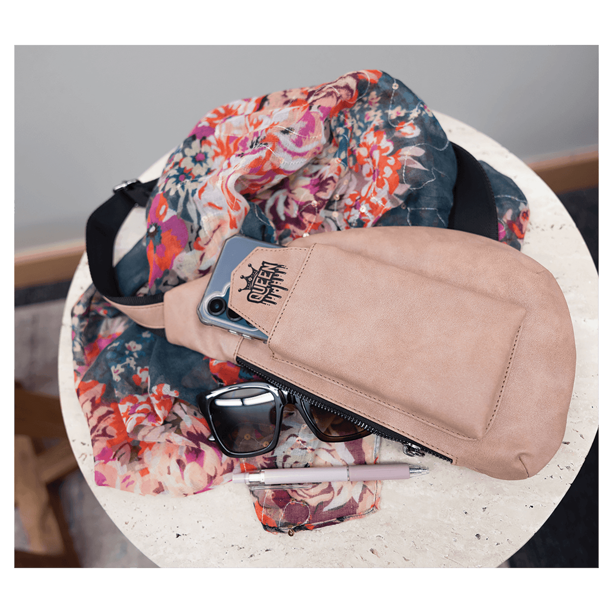 A beige crossbody bag holding a phone sits on a round white table next to a floral scarf, sunglasses, and a pen.