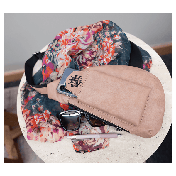 A beige crossbody bag holding a phone sits on a round white table next to a floral scarf, sunglasses, and a pen.
