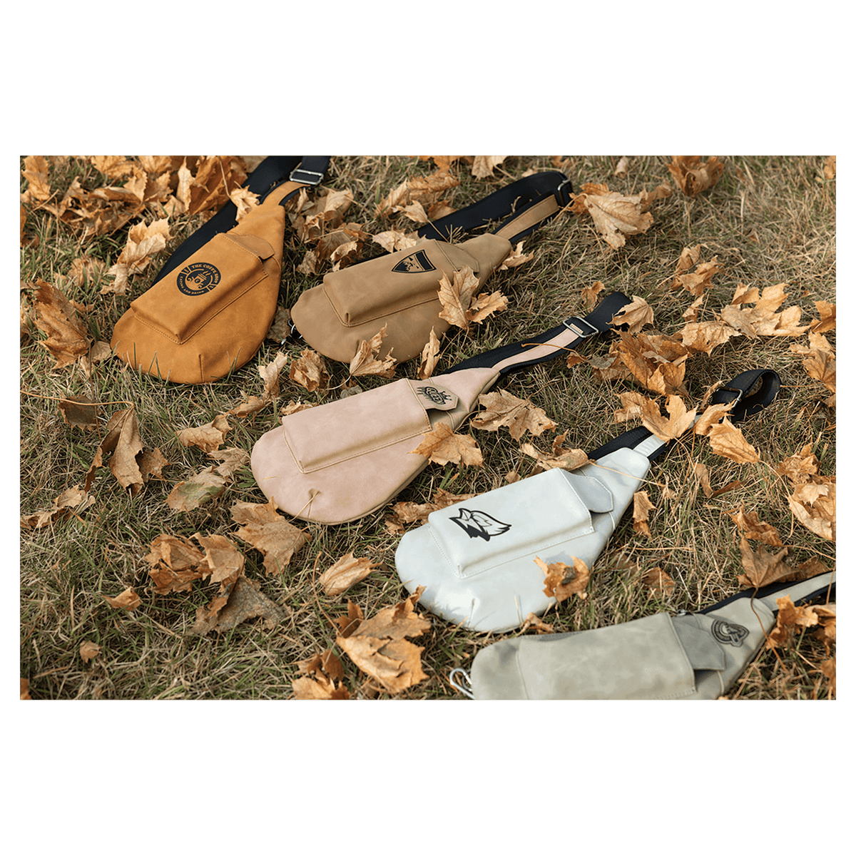 Five crossbody bags in orange, brown, beige, white, and grey lying flat on dry grass and fallen autumn leaves.
