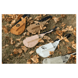 Five crossbody bags in orange, brown, beige, white, and grey lying flat on dry grass and fallen autumn leaves.
