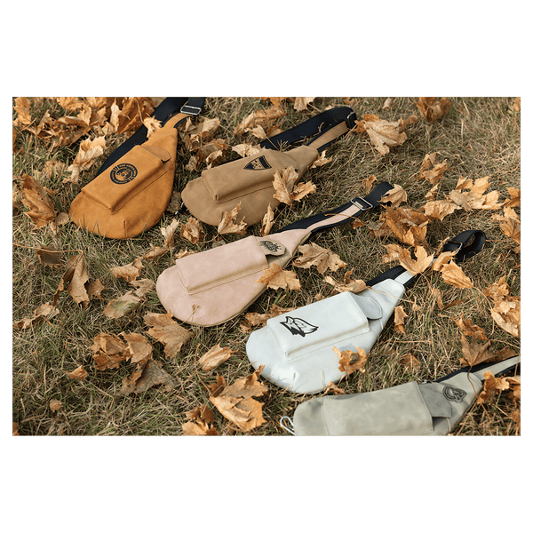 Five crossbody bags in orange, brown, beige, white, and grey lying flat on dry grass and fallen autumn leaves.