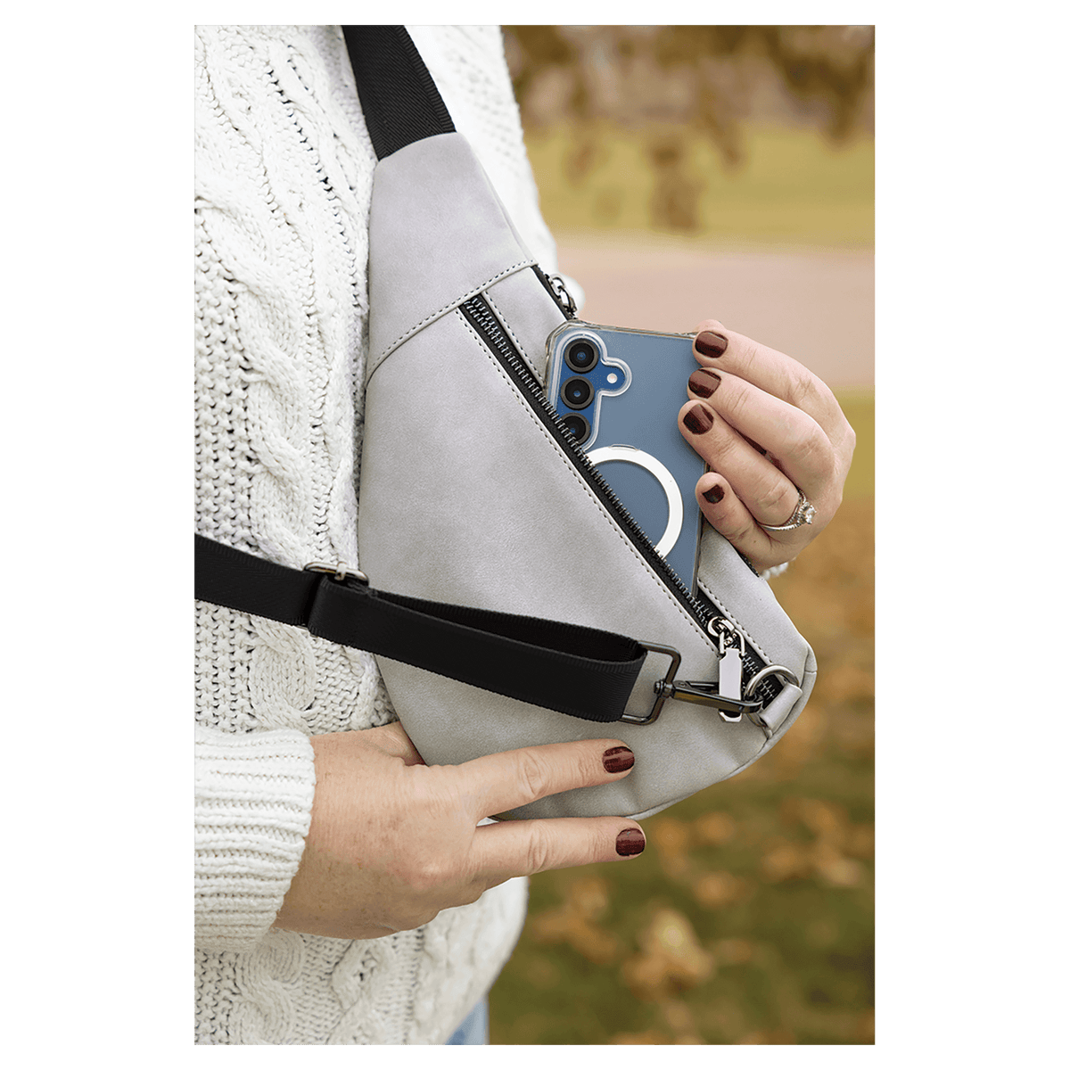 Person in a white sweater pulls a blue phone from a light grey crossbody bag against an autumn background.