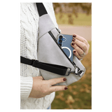 Person in a white sweater pulls a blue phone from a light grey crossbody bag against an autumn background.