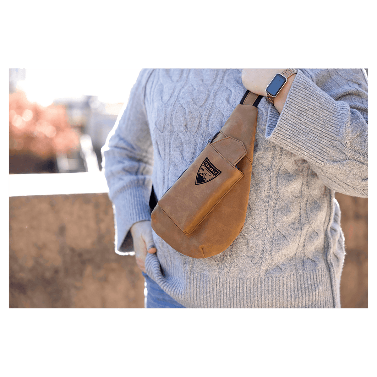 Person in a chunky grey sweater wears a brown crossbody sling bag outdoors in bright sunlight.