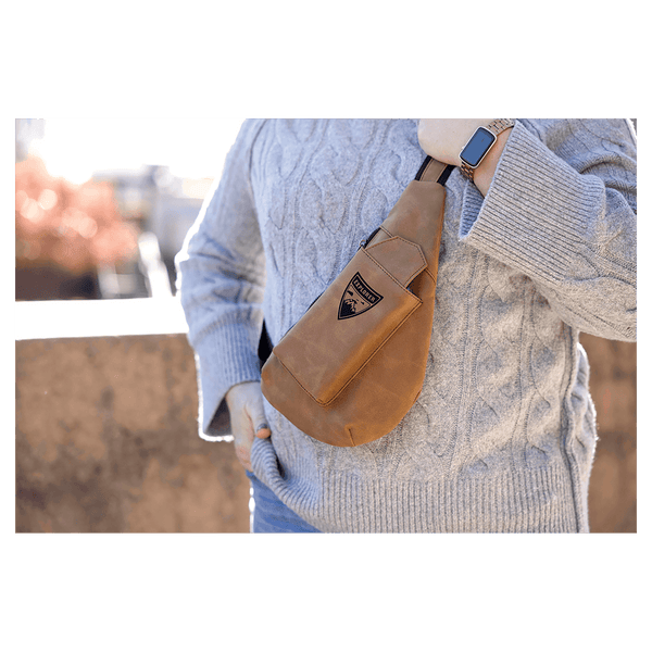 Person in a chunky grey sweater wears a brown crossbody sling bag outdoors in bright sunlight.