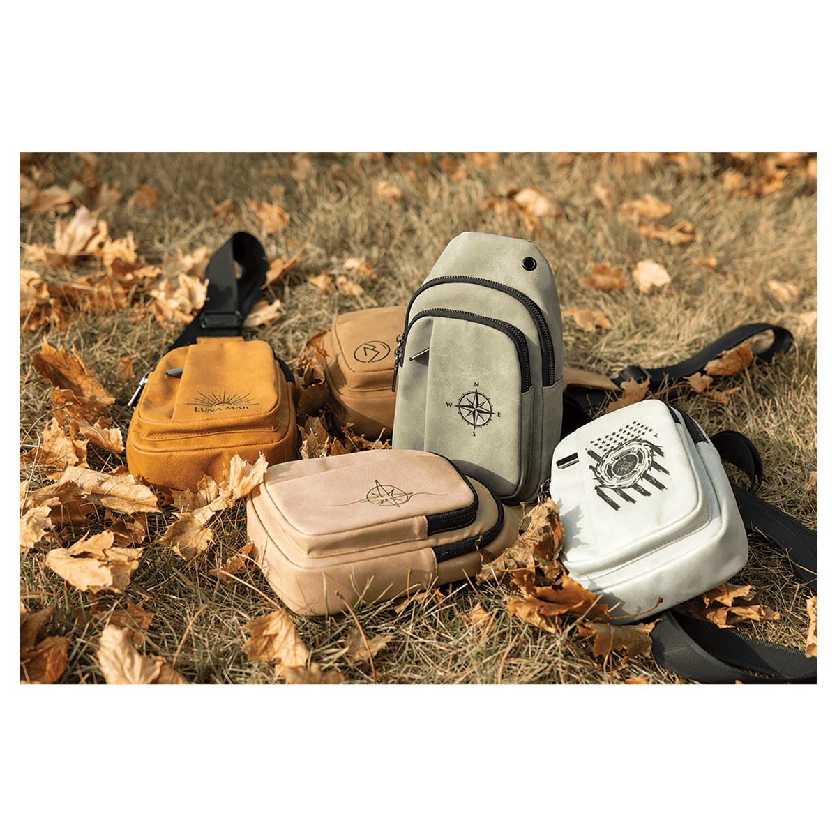 Five small crossbody bags in yellow, brown, gray, tan, and white with black straps scattered on dry autumn grass.