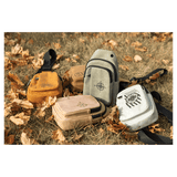 Five small crossbody bags in yellow, brown, gray, tan, and white with black straps scattered on dry autumn grass.