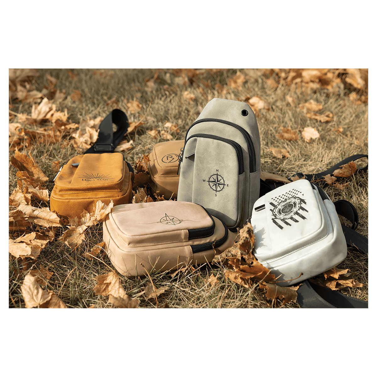 Four small sling bags in yellow, tan, grey, and white with black graphics, resting on dry grass and autumn leaves.