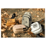 Four small sling bags in yellow, tan, grey, and white with black graphics, resting on dry grass and autumn leaves.