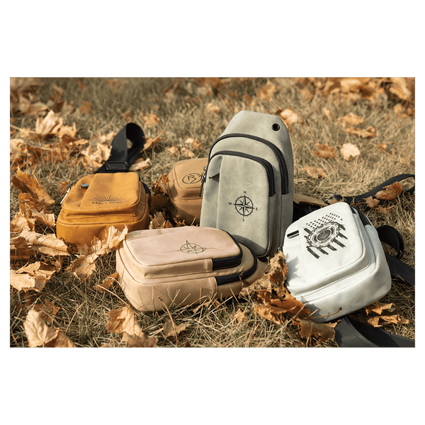 Four small sling bags in yellow, tan, grey, and white with black graphics, resting on dry grass and autumn leaves.