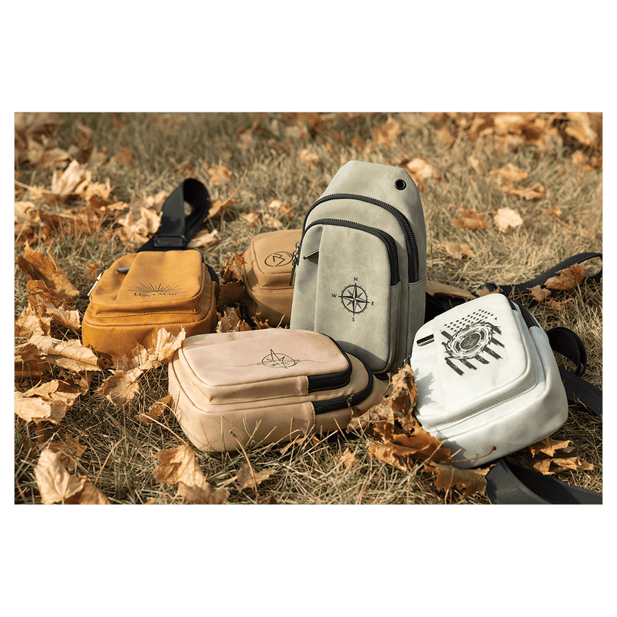 Four small sling bags in yellow, tan, grey, and white with black graphics, resting on dry grass and autumn leaves.