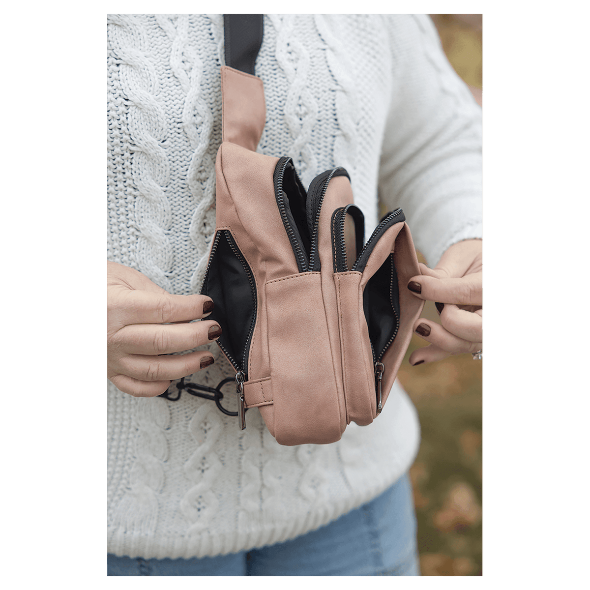 Person in a white sweater opens multiple zippered compartments of a pink crossbody bag against an autumn background.