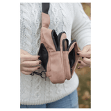 Person in a white sweater opens multiple zippered compartments of a pink crossbody bag against an autumn background.