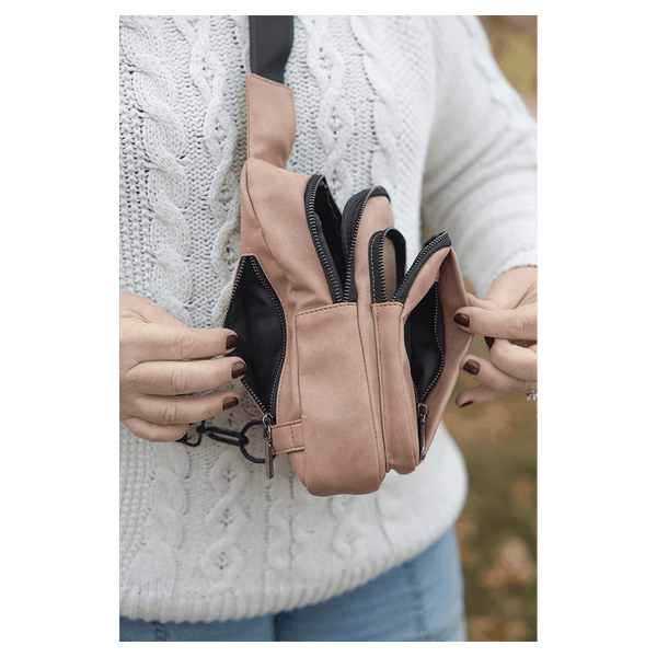 Person in a white sweater opens multiple zippered compartments of a pink crossbody bag against an autumn background.