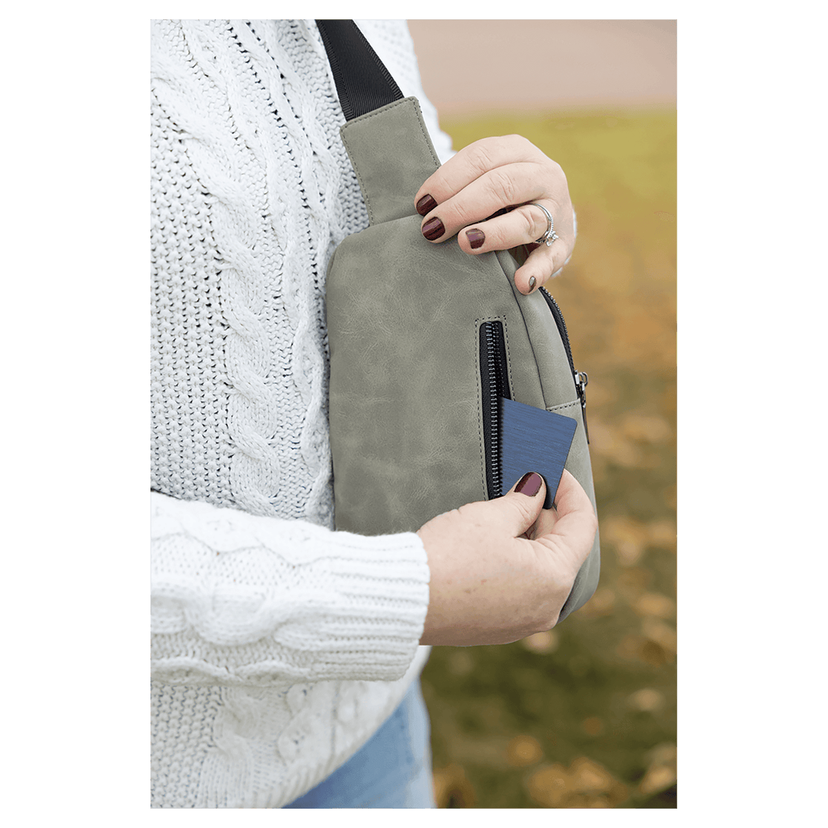 Person in a white sweater pulling a blue card from the back zipper pocket of a light gray sling bag outdoors.
