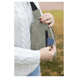 Person in a white sweater pulling a blue card from the back zipper pocket of a light gray sling bag outdoors.