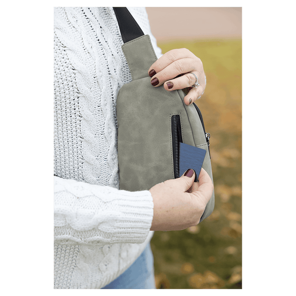Person in a white sweater pulling a blue card from the back zipper pocket of a light gray sling bag outdoors.
