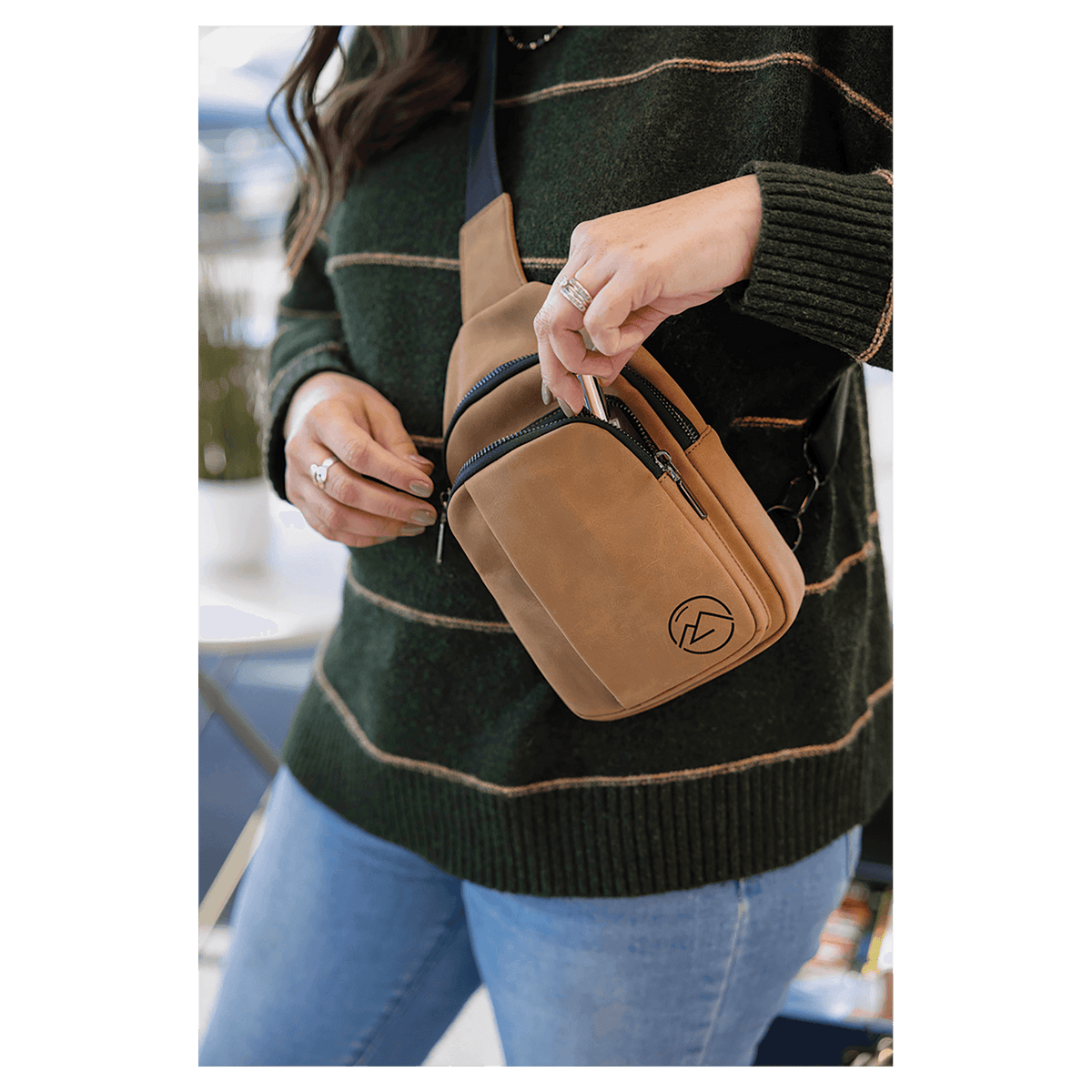 A person in a green striped sweater reaches into the zippered front pocket of a tan sling bag worn across the chest.