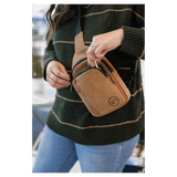 A person in a green striped sweater reaches into the zippered front pocket of a tan sling bag worn across the chest.