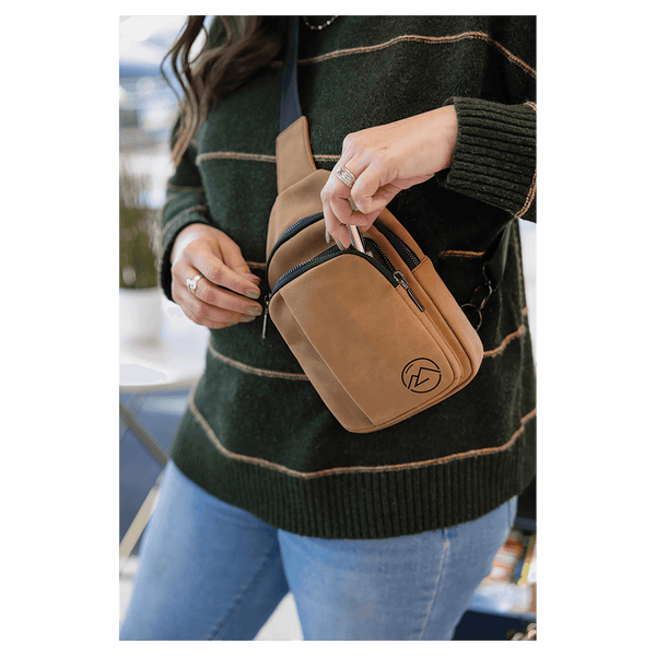 A person in a green striped sweater reaches into the zippered front pocket of a tan sling bag worn across the chest.