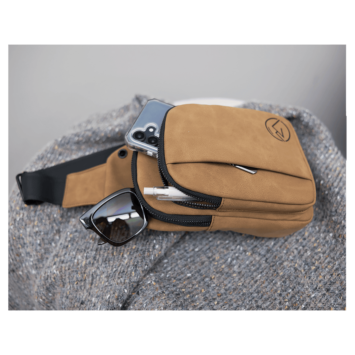 Tan crossbody bag rests on a gray knit blanket, holding a phone, sunglasses, and pen in its open zippered pocket.