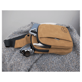 Tan crossbody bag rests on a gray knit blanket, holding a phone, sunglasses, and pen in its open zippered pocket.