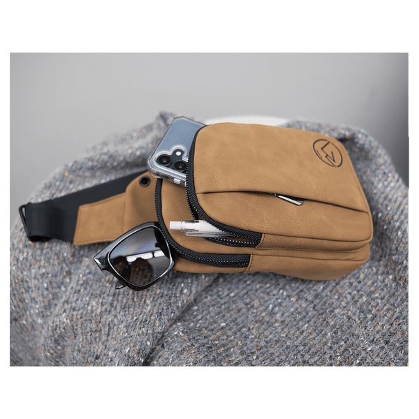 Tan crossbody bag rests on a gray knit blanket, holding a phone, sunglasses, and pen in its open zippered pocket.