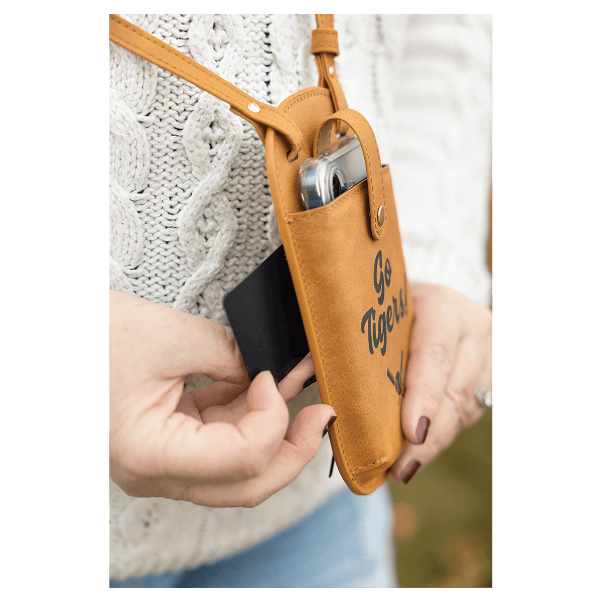 Hands pull a card from a brown crossbody phone bag printed with "Go Tigers!" against a white sweater and blue jeans.