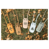 Five personalized phone bags in yellow, brown, beige, blue, and green resting on grass with autumn leaves.