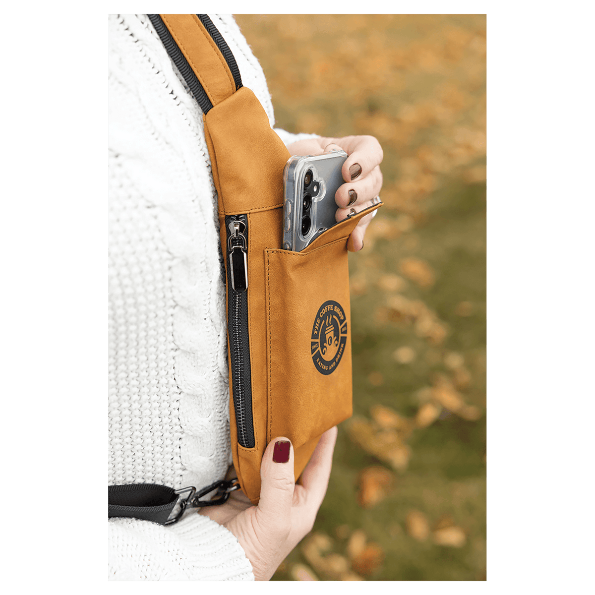 A person in a white sweater removes a phone from a tan crossbody bag front pocket outdoors against autumn leaves.