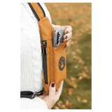 A person in a white sweater removes a phone from a tan crossbody bag front pocket outdoors against autumn leaves.