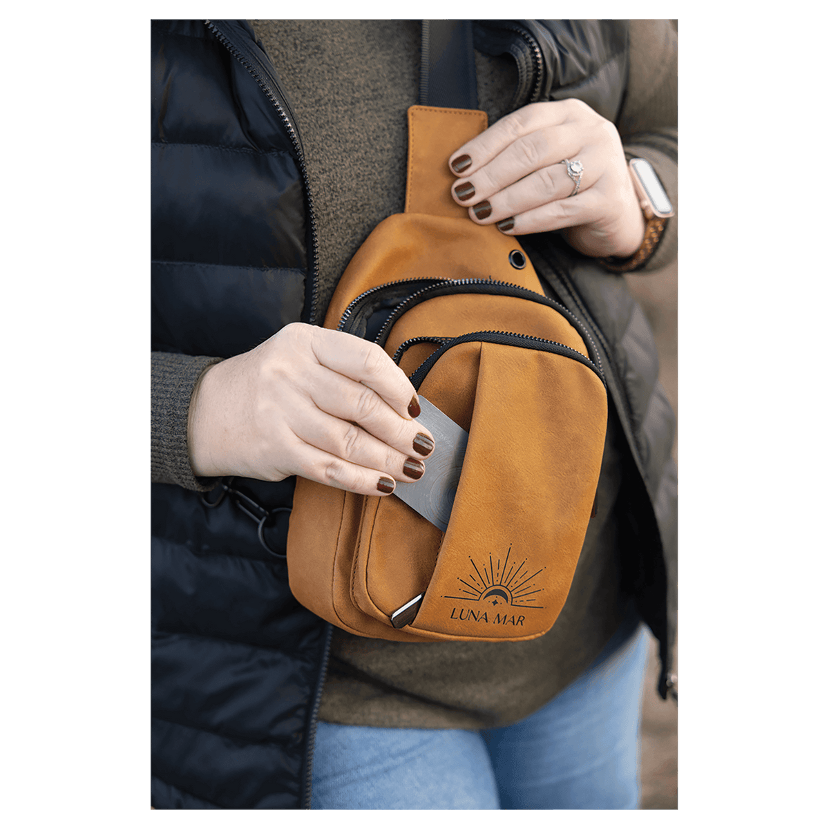 Person in a black vest wearing a mustard yellow sling bag, taking a silver card out of its front slip pocket.