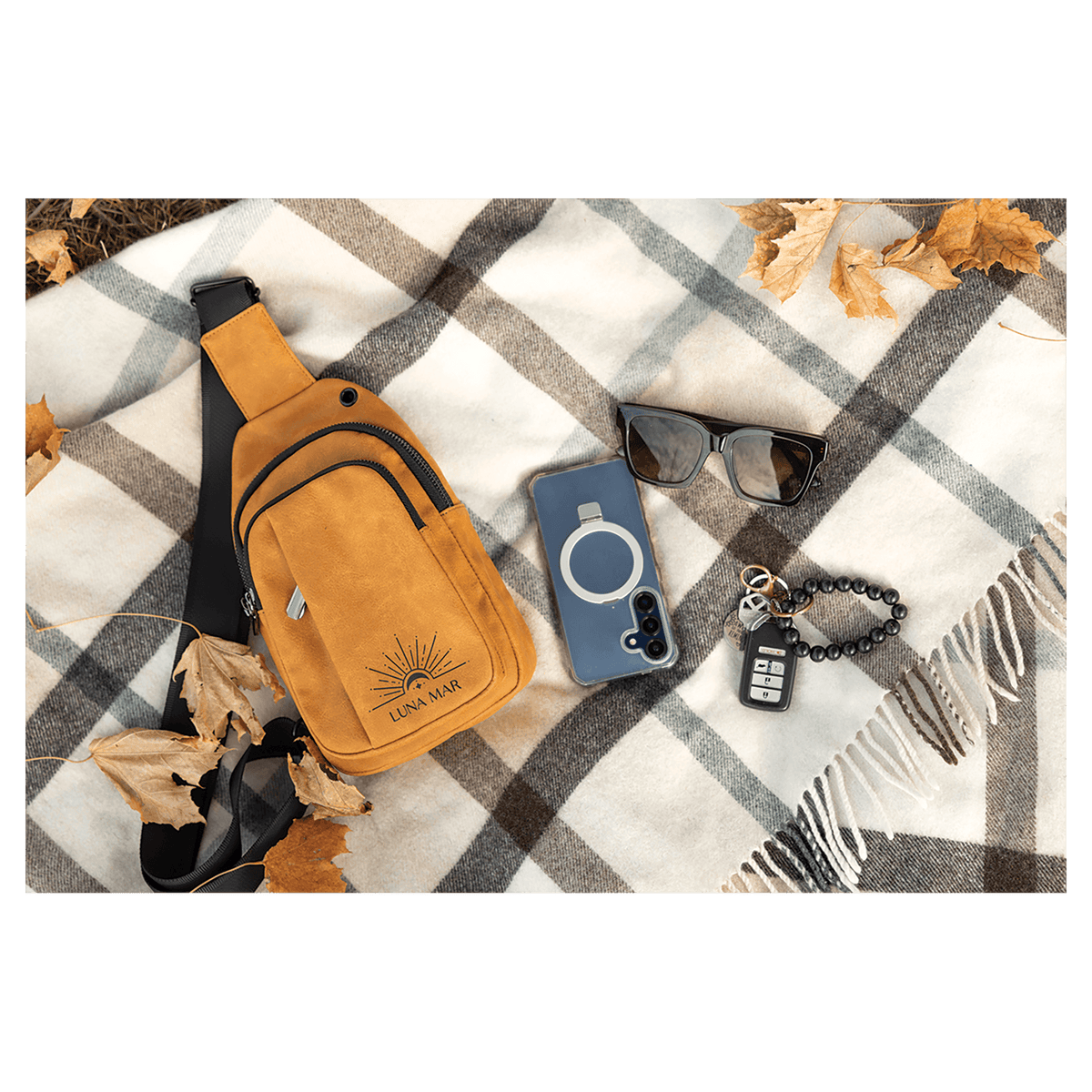 Mustard yellow sling bag, blue phone, black sunglasses, and keys flat on a grey and white plaid blanket with leaves.