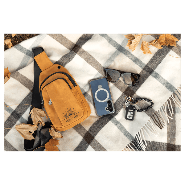 Mustard yellow sling bag, blue phone, black sunglasses, and keys flat on a grey and white plaid blanket with leaves.
