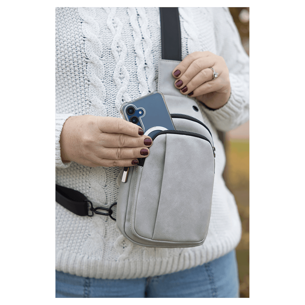 Person wearing a white sweater taking a blue smartphone out of a light gray crossbody bag worn on their chest.