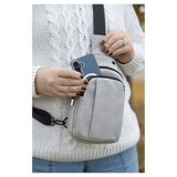 Person wearing a white sweater taking a blue smartphone out of a light gray crossbody bag worn on their chest.