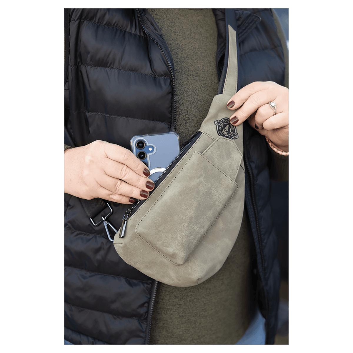 Person wearing a black vest pulls a blue smartphone from an olive green crossbody bag.