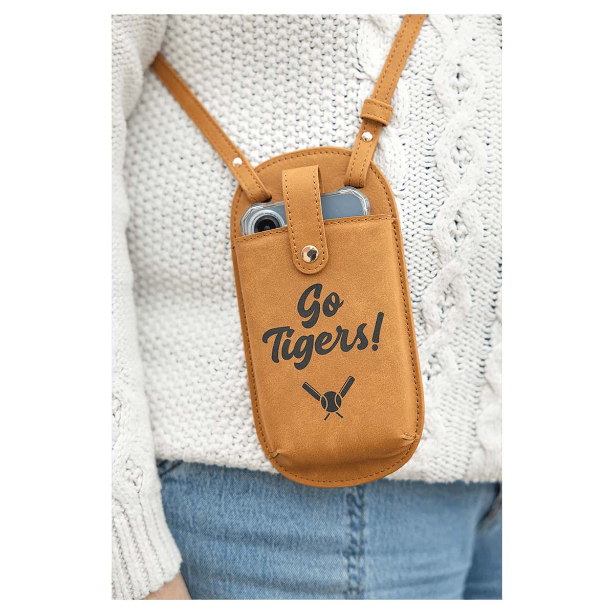 A brown crossbody phone bag printed with "Go Tigers!" and baseball bats, worn over a white sweater and blue jeans.