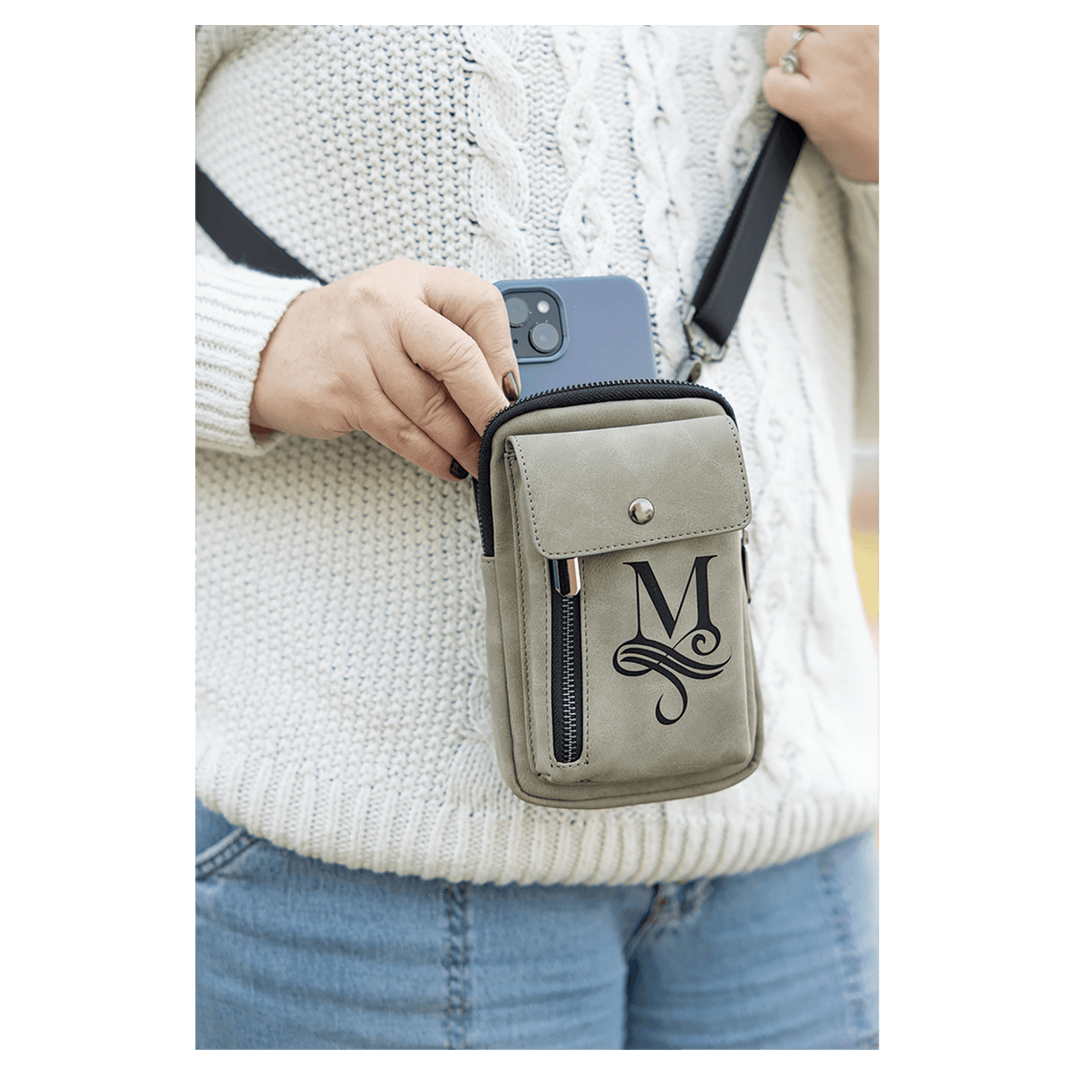 Taupe crossbody bag with a black strap, front snap flap, and vertical zip, worn over a white sweater holding a phone.