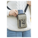 Taupe crossbody bag with a black strap, front snap flap, and vertical zip, worn over a white sweater holding a phone.