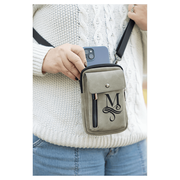 Taupe crossbody bag with a black strap, front snap flap, and vertical zip, worn over a white sweater holding a phone.