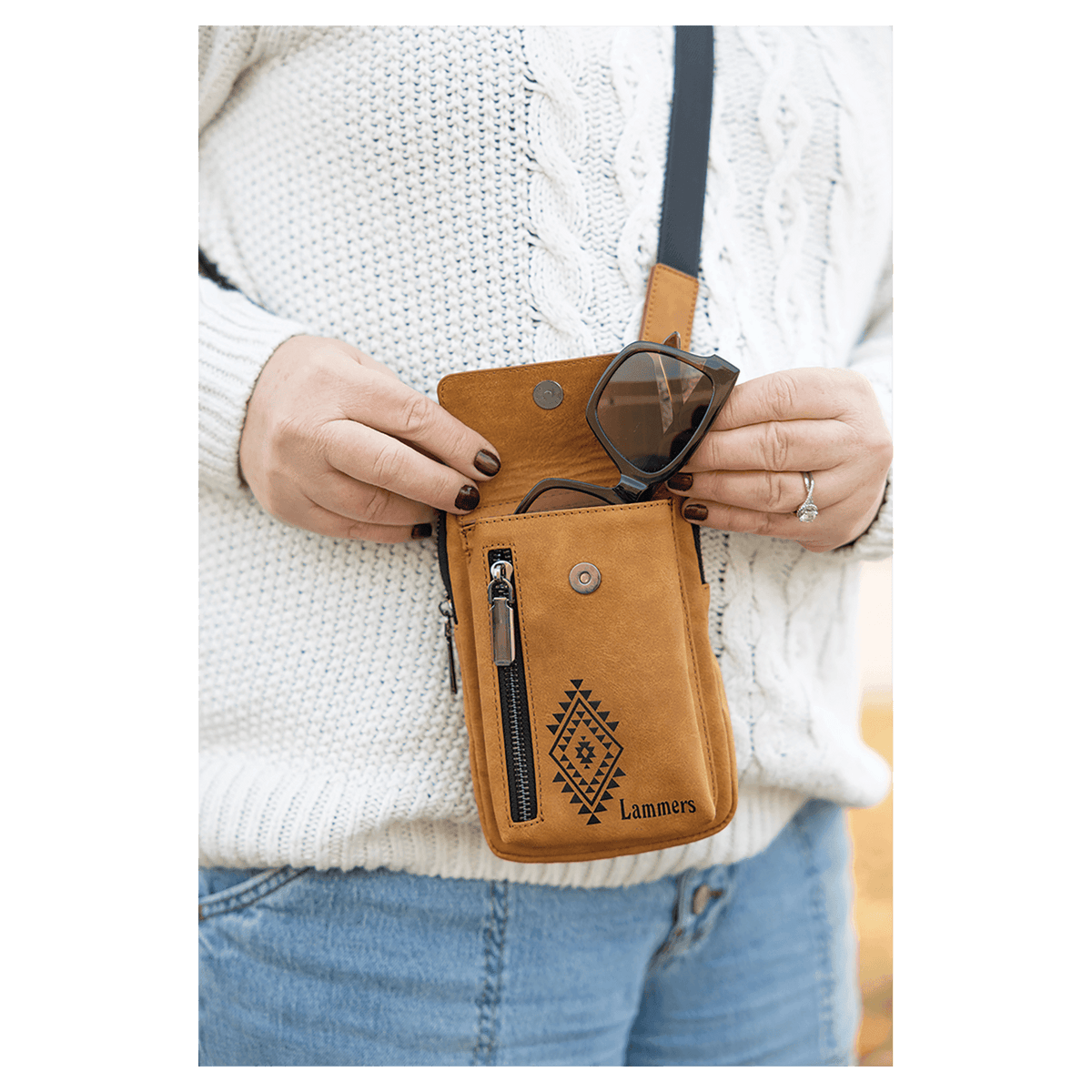 Tan crossbody bag with a vertical zipper and black strap, worn over a white sweater with sunglasses in the open flap.