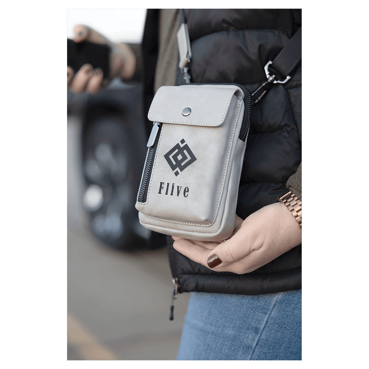 Light grey crossbody bag with a front snap flap, vertical zipper, and black strap, worn over a black puffer vest.