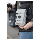 Light grey crossbody bag with a front snap flap, vertical zipper, and black strap, worn over a black puffer vest.
